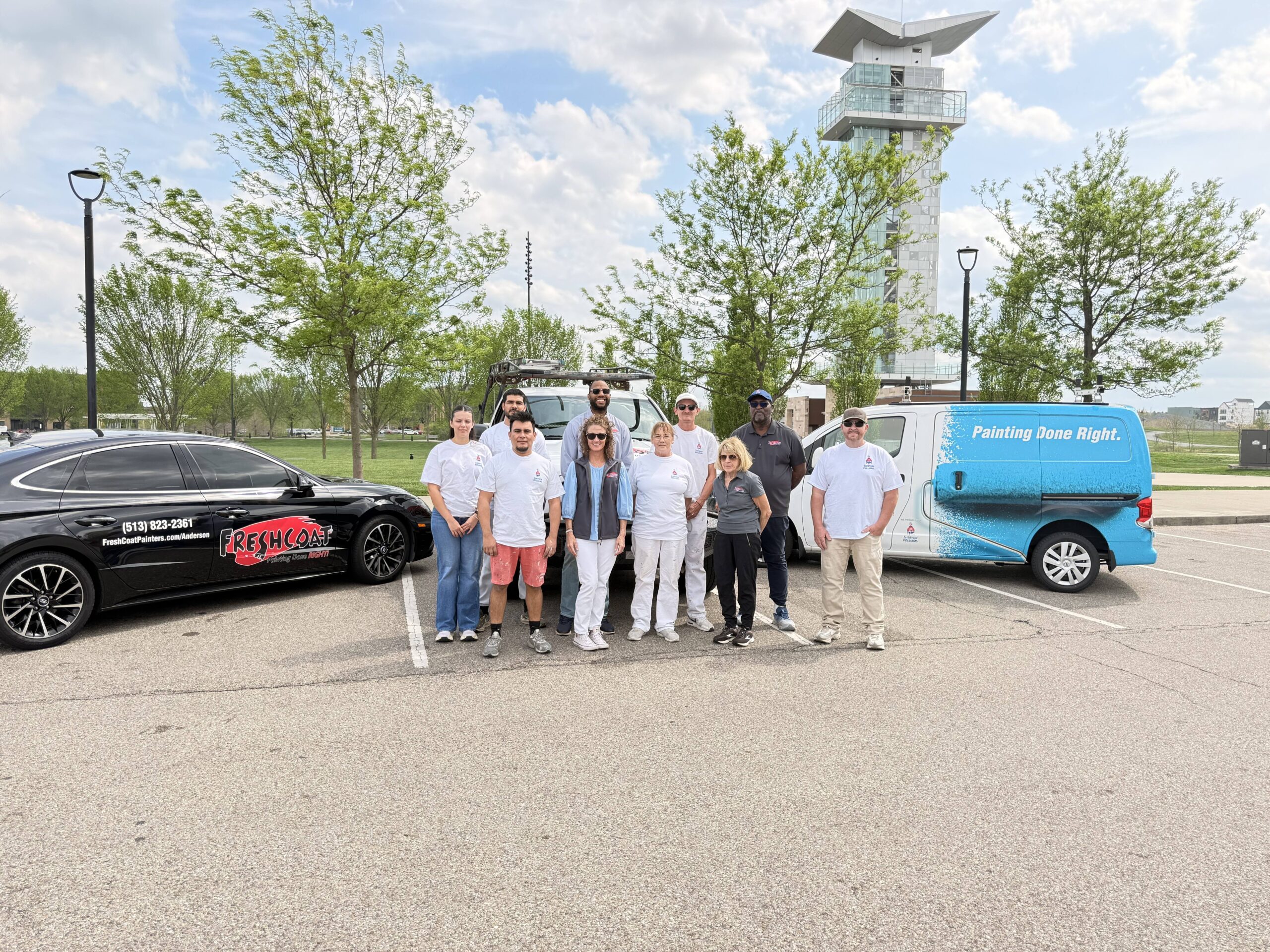 Group of painters posing in a parking lot between a black company car and a blue van that says 'Painting Done Right', with a tall tower in the background.